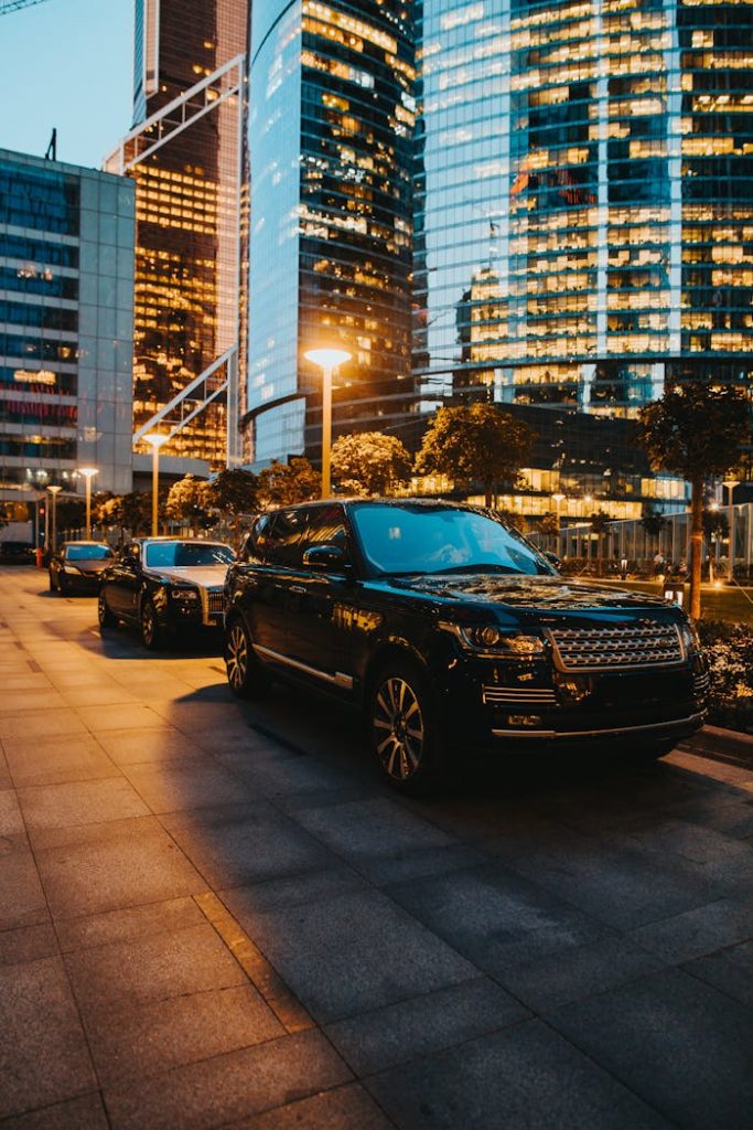pexels photo 5058352 Elegant SUVs parked along illuminated city streets at dusk, reflecting modern urban architecture.